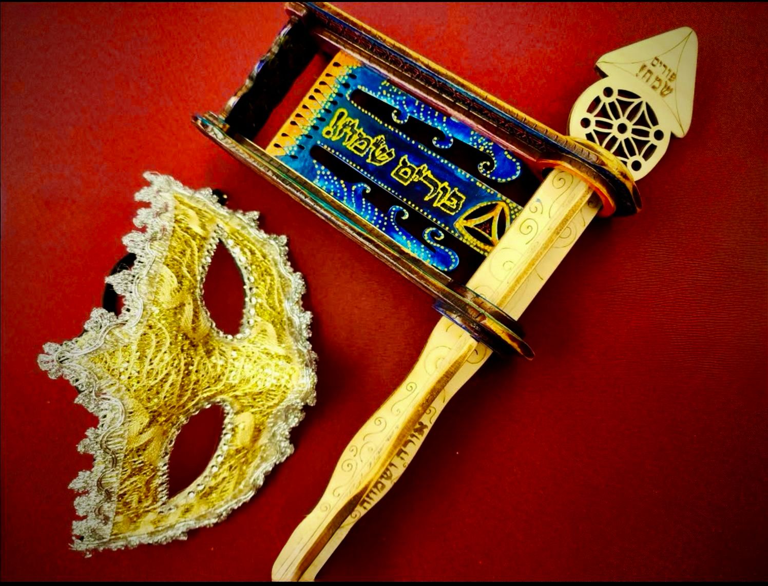 Intentional Purim Gragger flat-lay on red background with gold masquerade mask